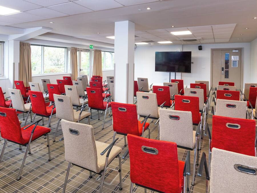 A conference room with rows of red and beige chairs, a flat-screen TV on the wall, and large windows with curtains.