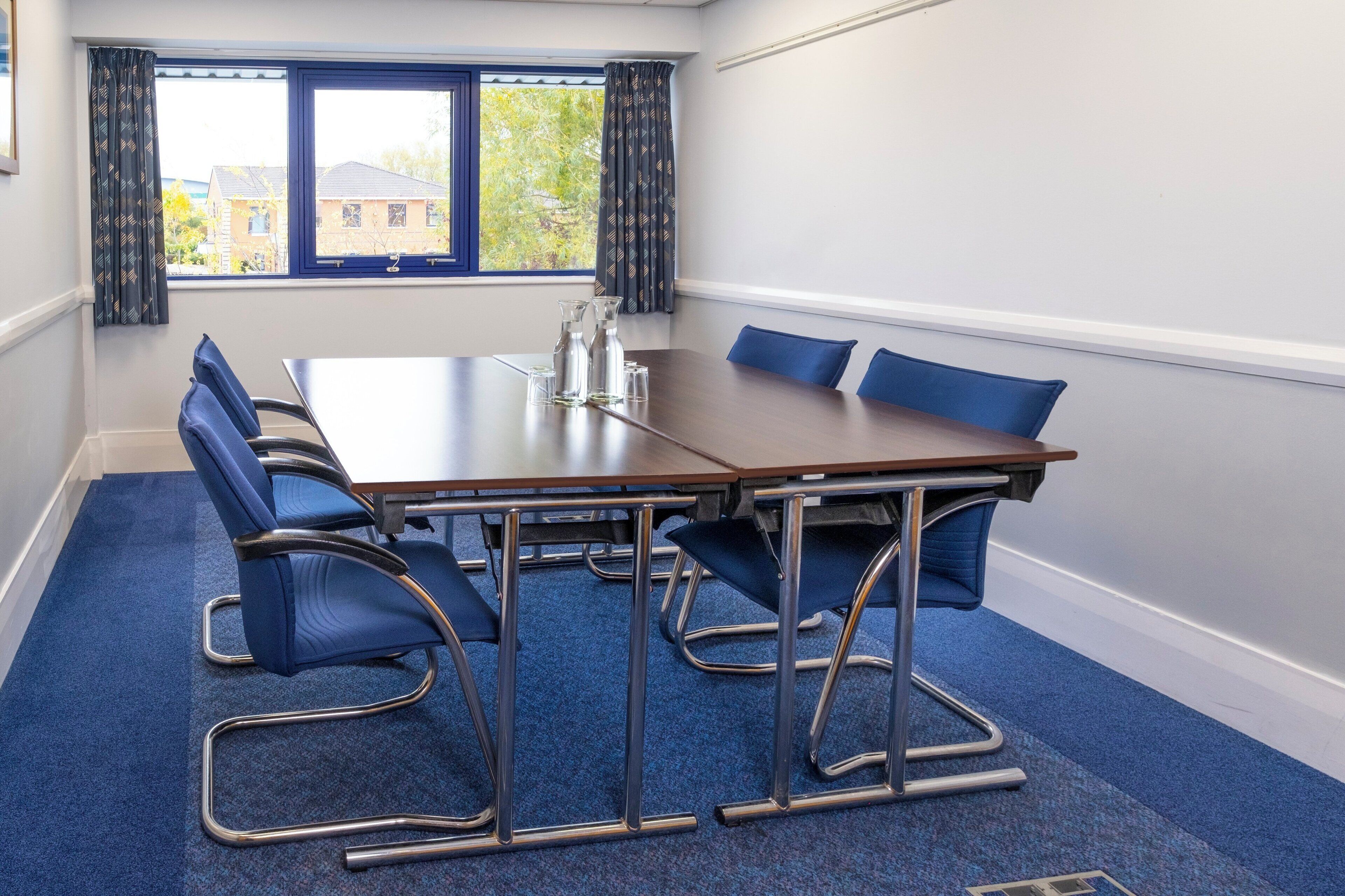 A conference room with a long table, six blue chairs, a pitcher, and glasses on the table.