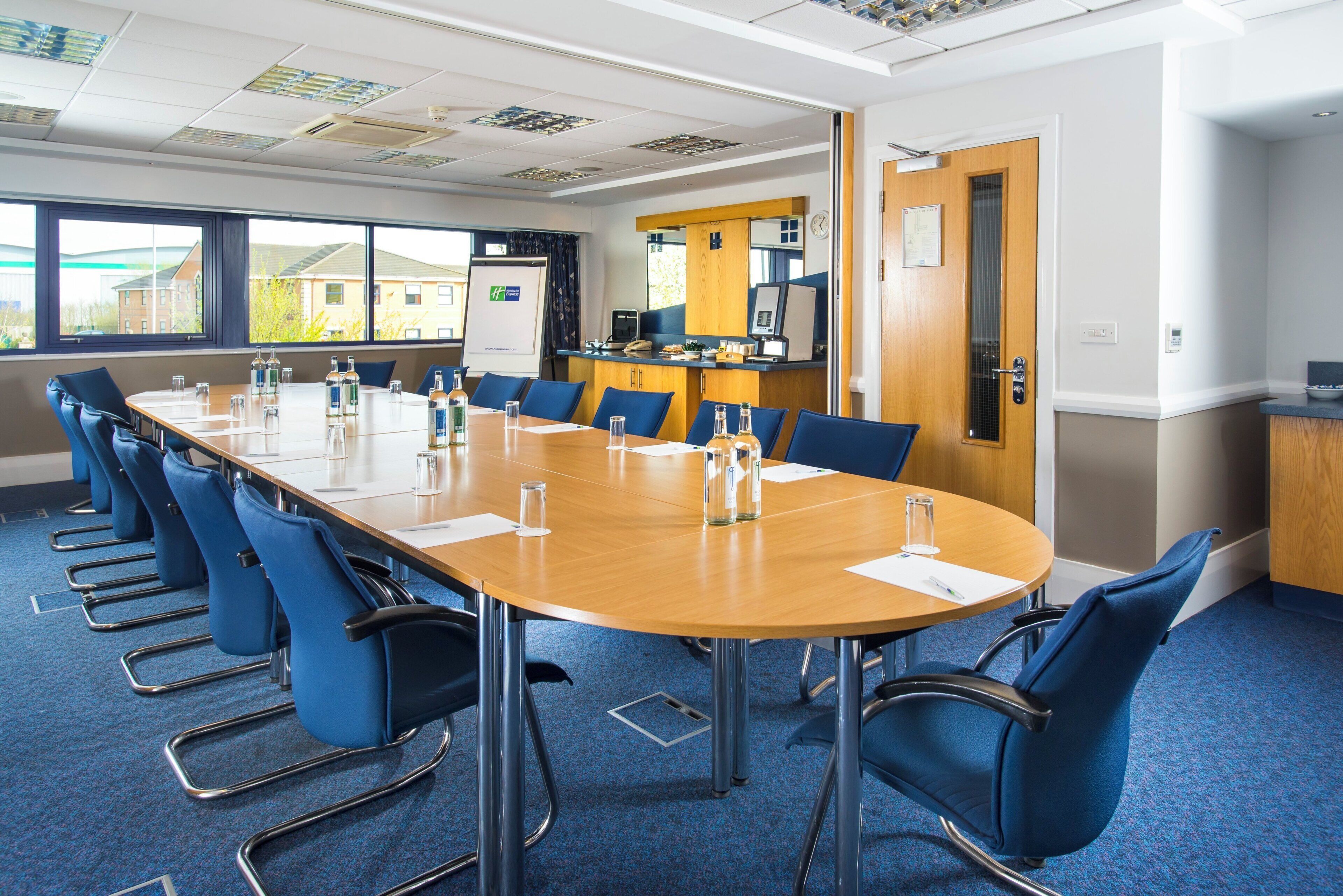 A conference room with a long table, blue chairs, and a kitchen area in the background.