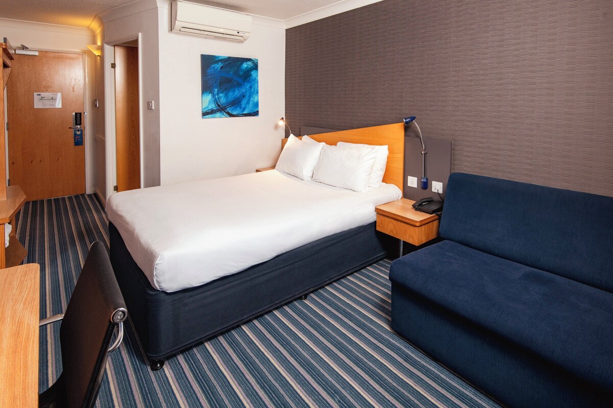 A hotel room with a bed, a bedside table with a lamp, a blue sofa, and a striped carpet.