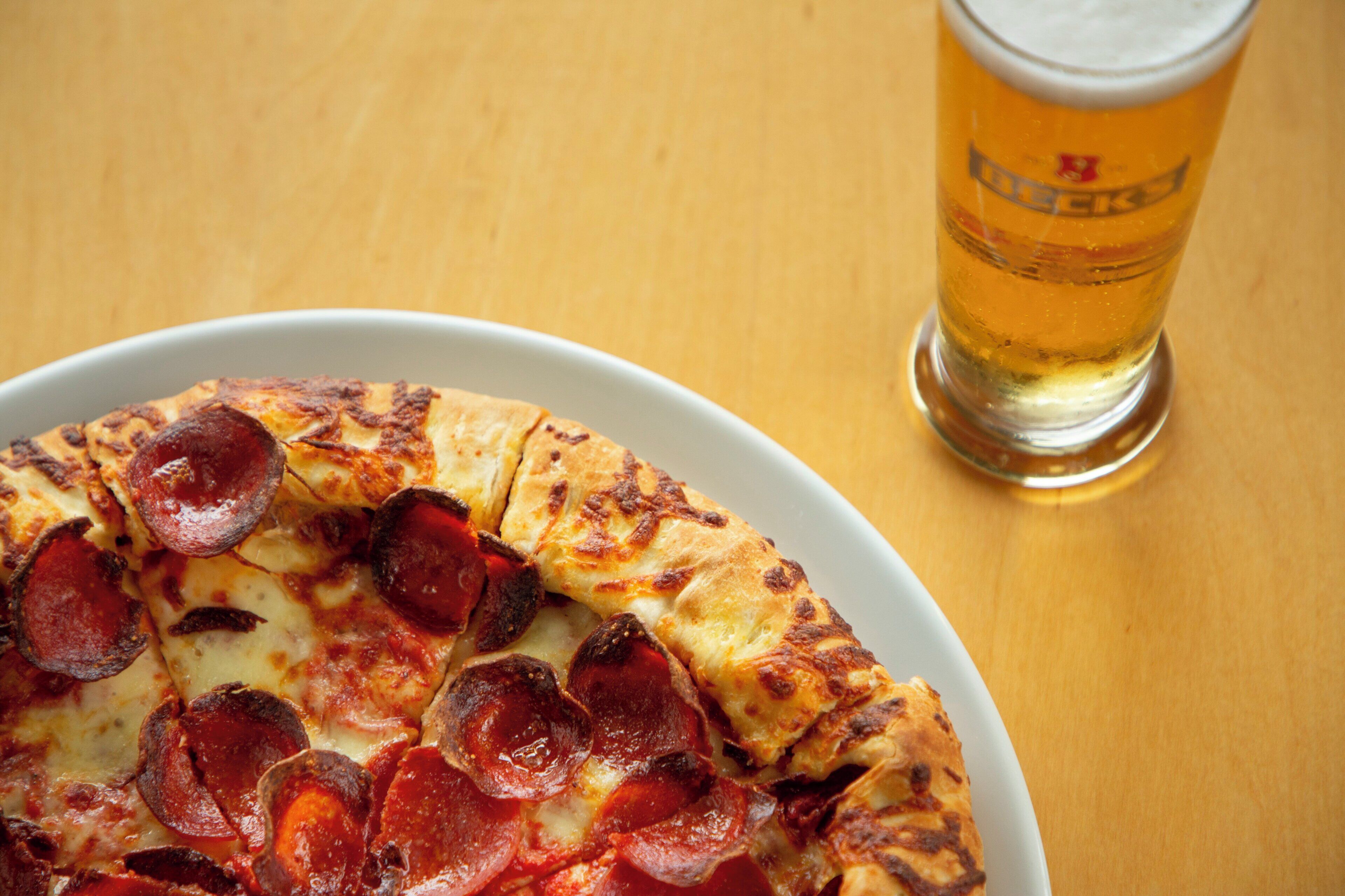Pizza con rodajas de pepperoni sobre un plato blanco, acompañada de una cerveza en una mesa de madera.