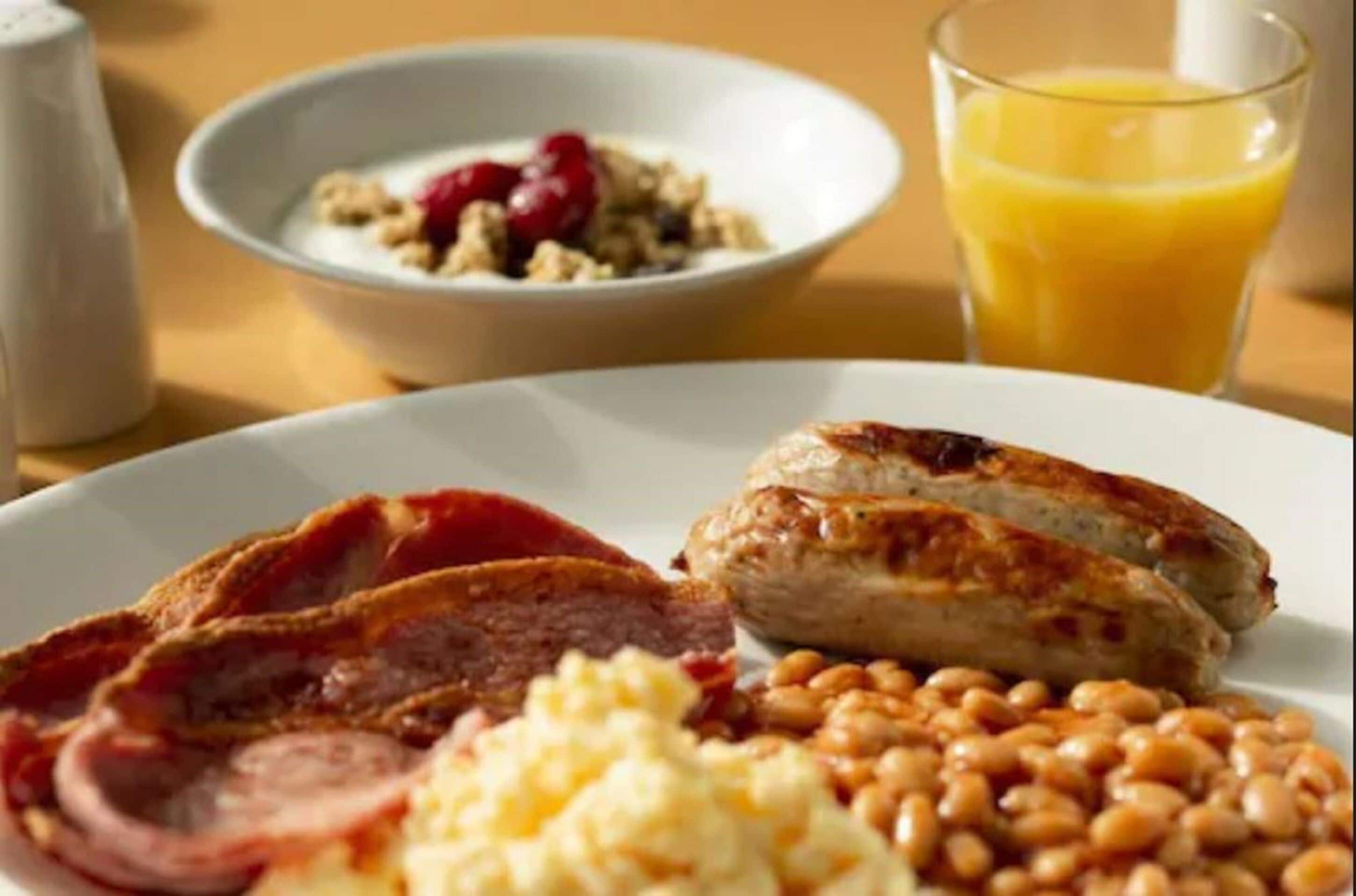 A plate with bacon, sausages, beans, scrambled eggs, and a bowl of cereal with fruit.