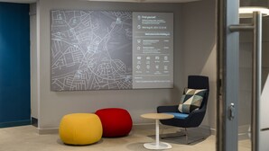 A modern hotel lobby with a large map on the wall, a seating area with a round table, and two colorful ottomans. - Holiday Inn Express London - Park Royal by IHG (London)