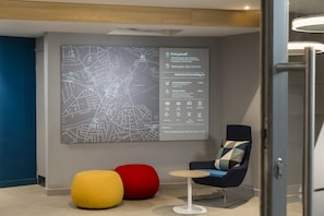 A modern hotel lobby with a large map on the wall, a seating area with a round table, and two colorful ottomans. - Holiday Inn Express London - Park Royal by IHG (London)