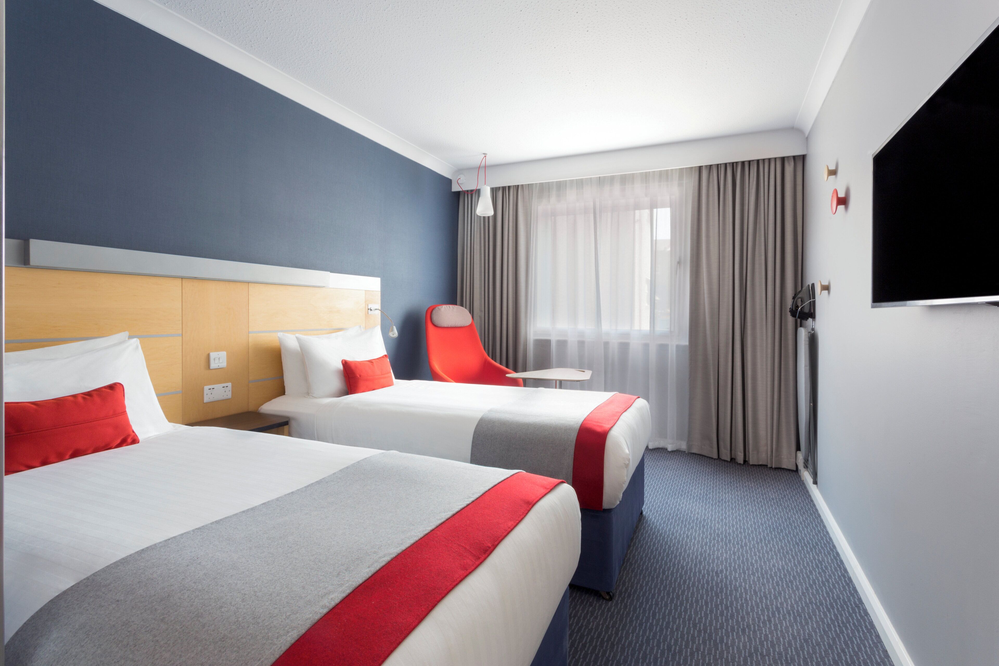 Une chambre d’hôtel avec deux lits, un fauteuil rouge, une télévision et une fenêtre avec des rideaux.