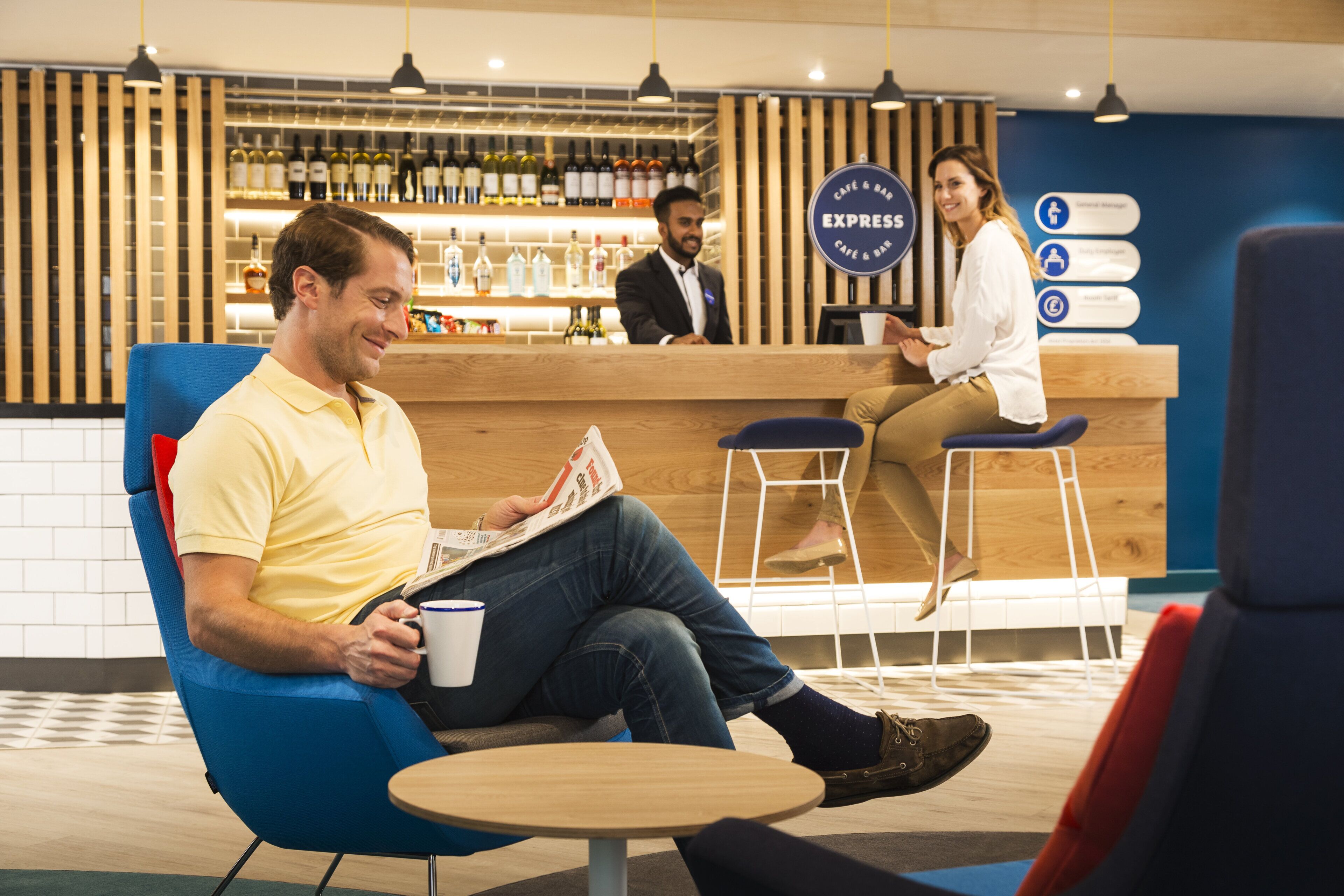 A man sitting on a blue chair reading a newspaper, with a bar and counter in the background.