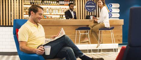 A man sitting on a blue chair reading a newspaper, with a bar and counter in the background.