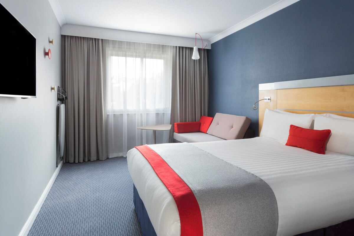 Une chambre d’hôtel avec un grand lit, une télévision, une petite table et une fenêtre avec des rideaux.