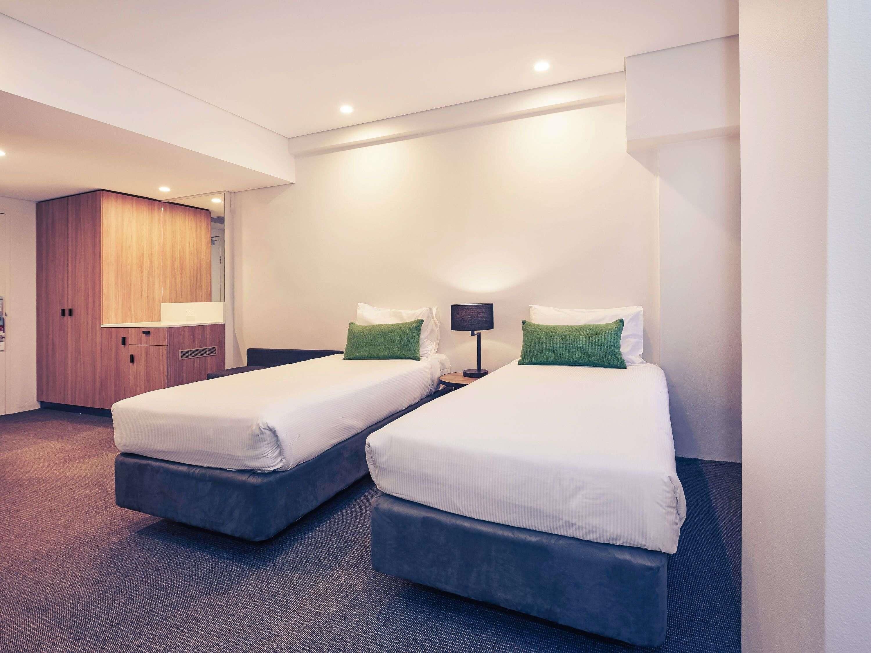 A hotel room with two beds, a wooden wardrobe, a nightstand with a lamp, and a carpeted floor.