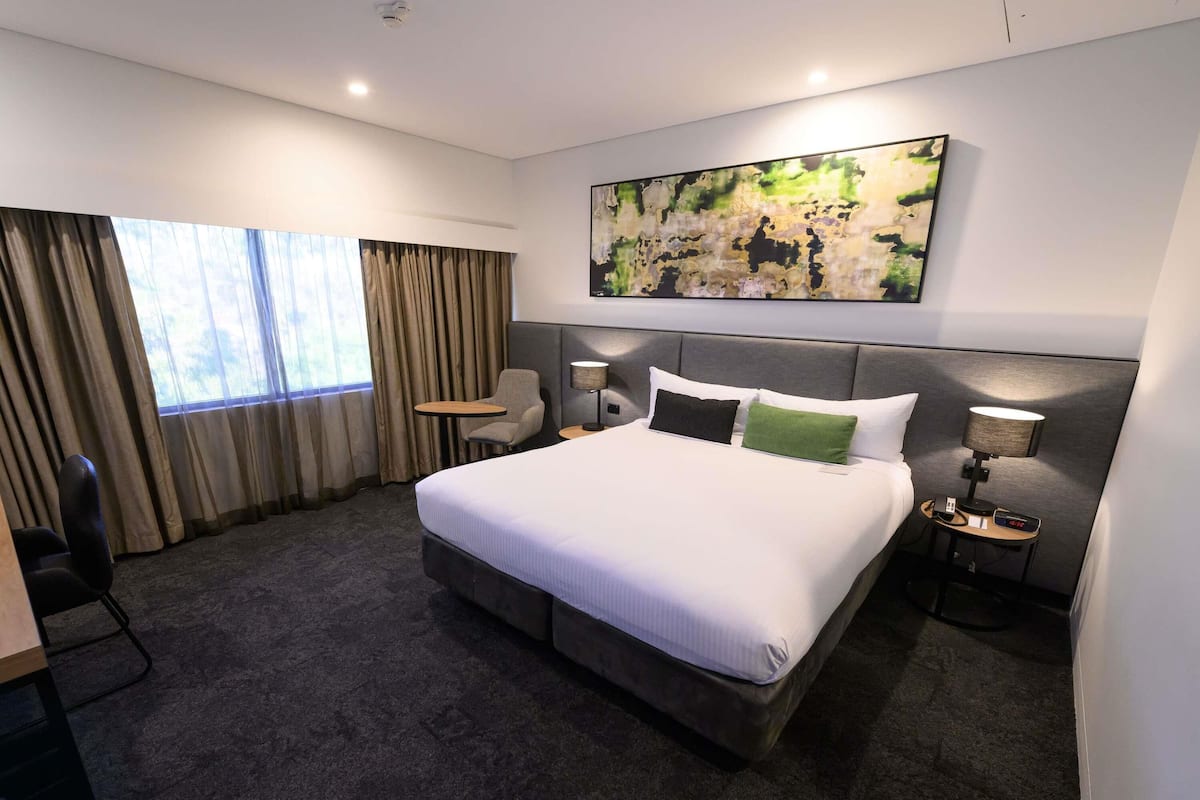Una habitación de hotel con una cama grande, un escritorio, una silla, una mesita, una lámpara y un cuadro en la pared.