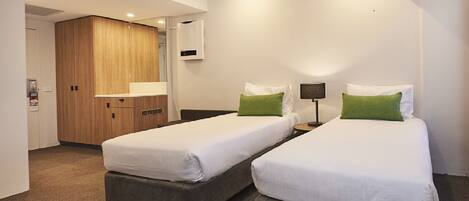 A hotel room with two beds, a wooden wardrobe, a small desk with a lamp, and a mirror on the wall.