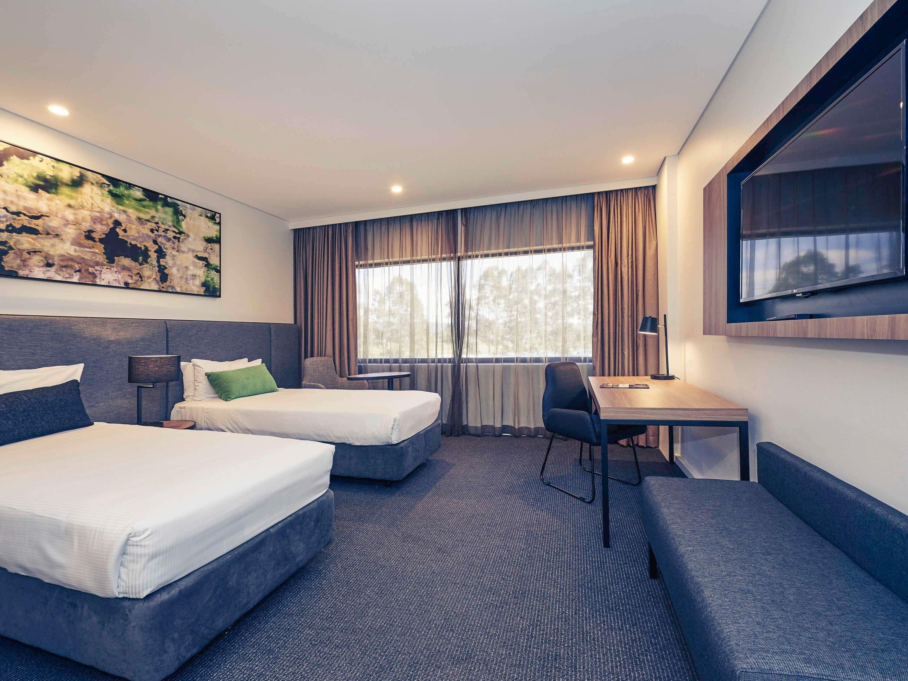 A hotel room with two beds, a desk, a chair, a TV, and a large window with curtains.