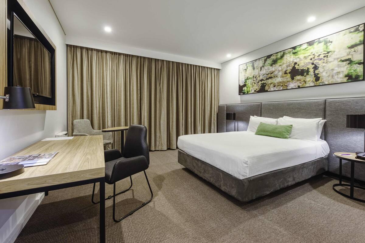 A hotel room with a bed, desk, chair, and a large abstract painting on the wall.