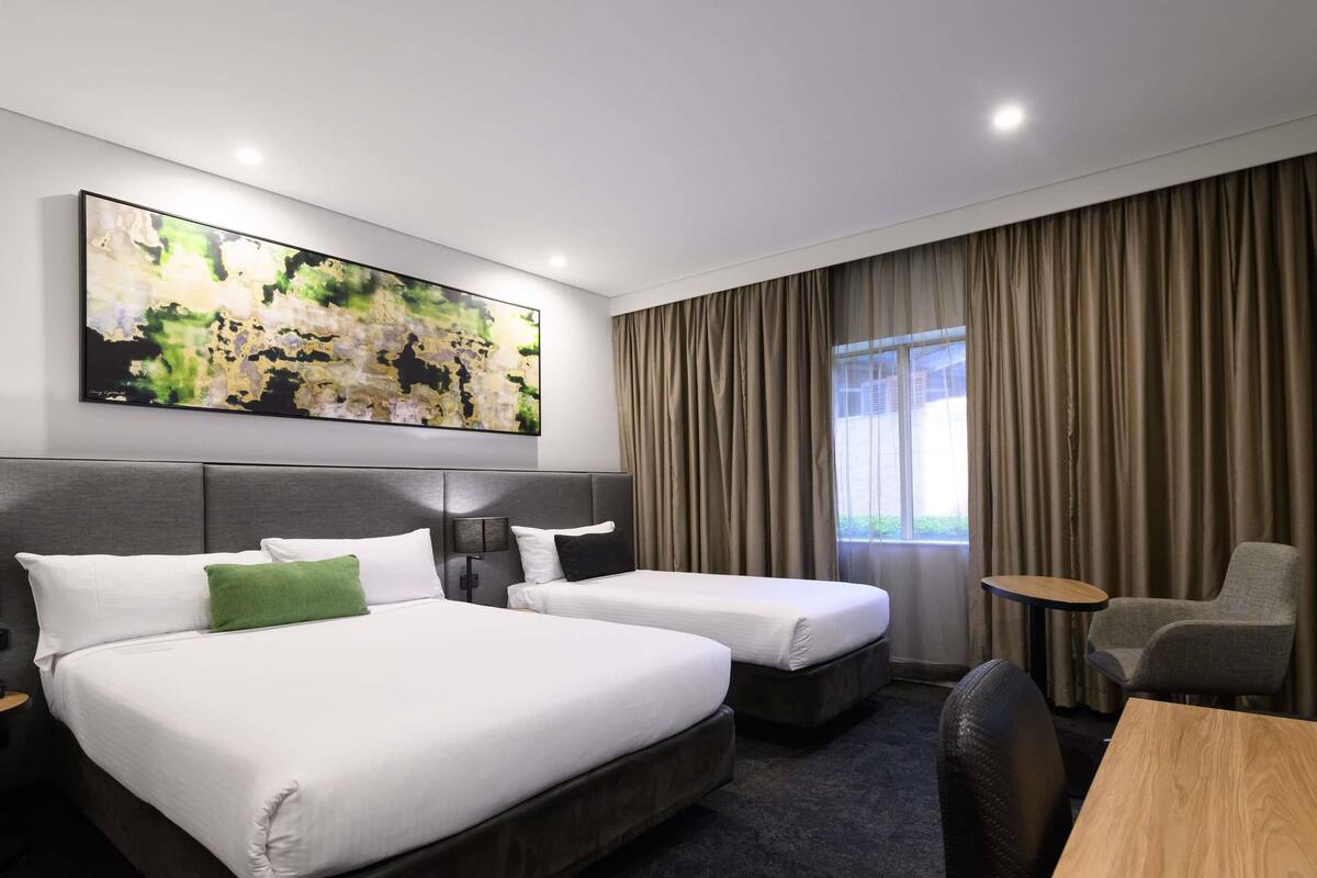 A hotel room with two beds, a desk, a chair, a large abstract painting, and a window with curtains.