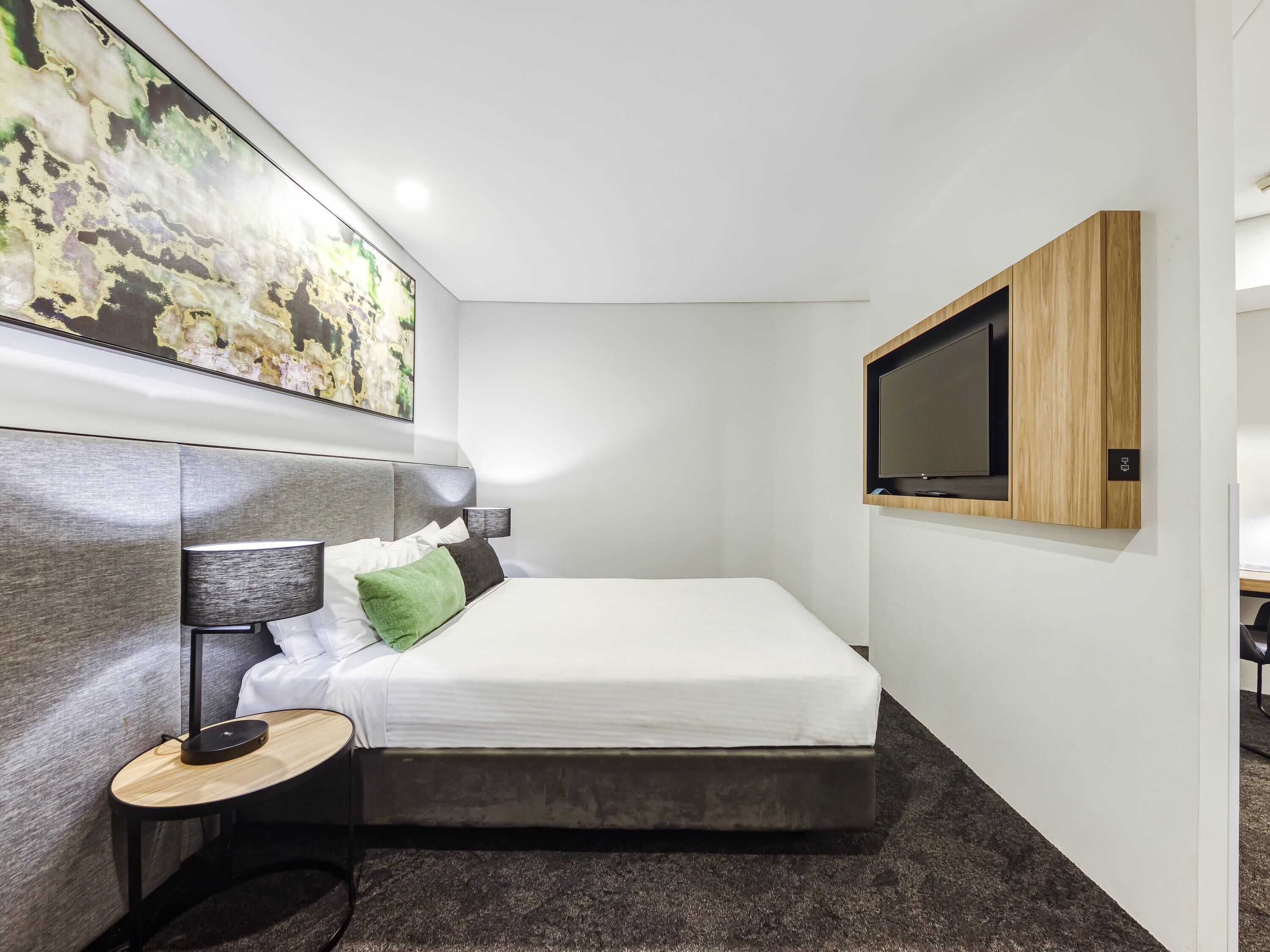 Ein modernes Hotelzimmer mit Bett, Nachttisch, Lampe, Kunstwerk und einem an der Wand befestigten Fernseher.