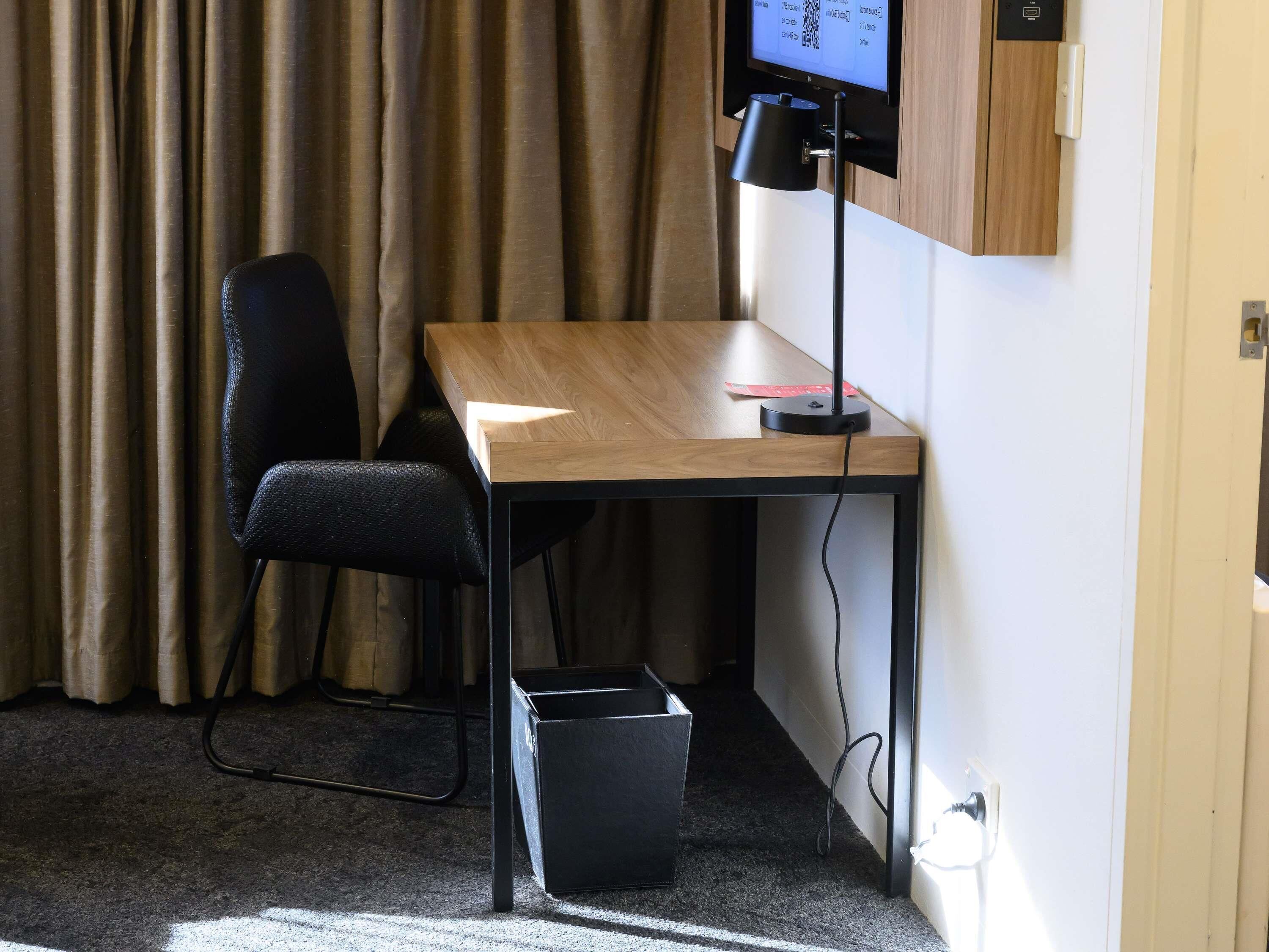 A hotel room with a wooden desk, a black office chair, a black trash bin, and a television mounted on the wall.