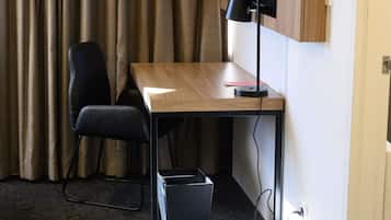 A hotel room with a wooden desk, a black office chair, a black trash bin, and a television mounted on the wall.