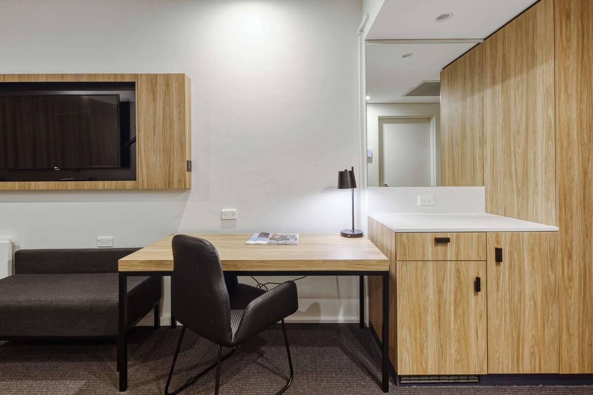 Una habitación moderna con un escritorio de madera, una silla negra, un televisor de pantalla plana montado en la pared y una encimera blanca con armarios.