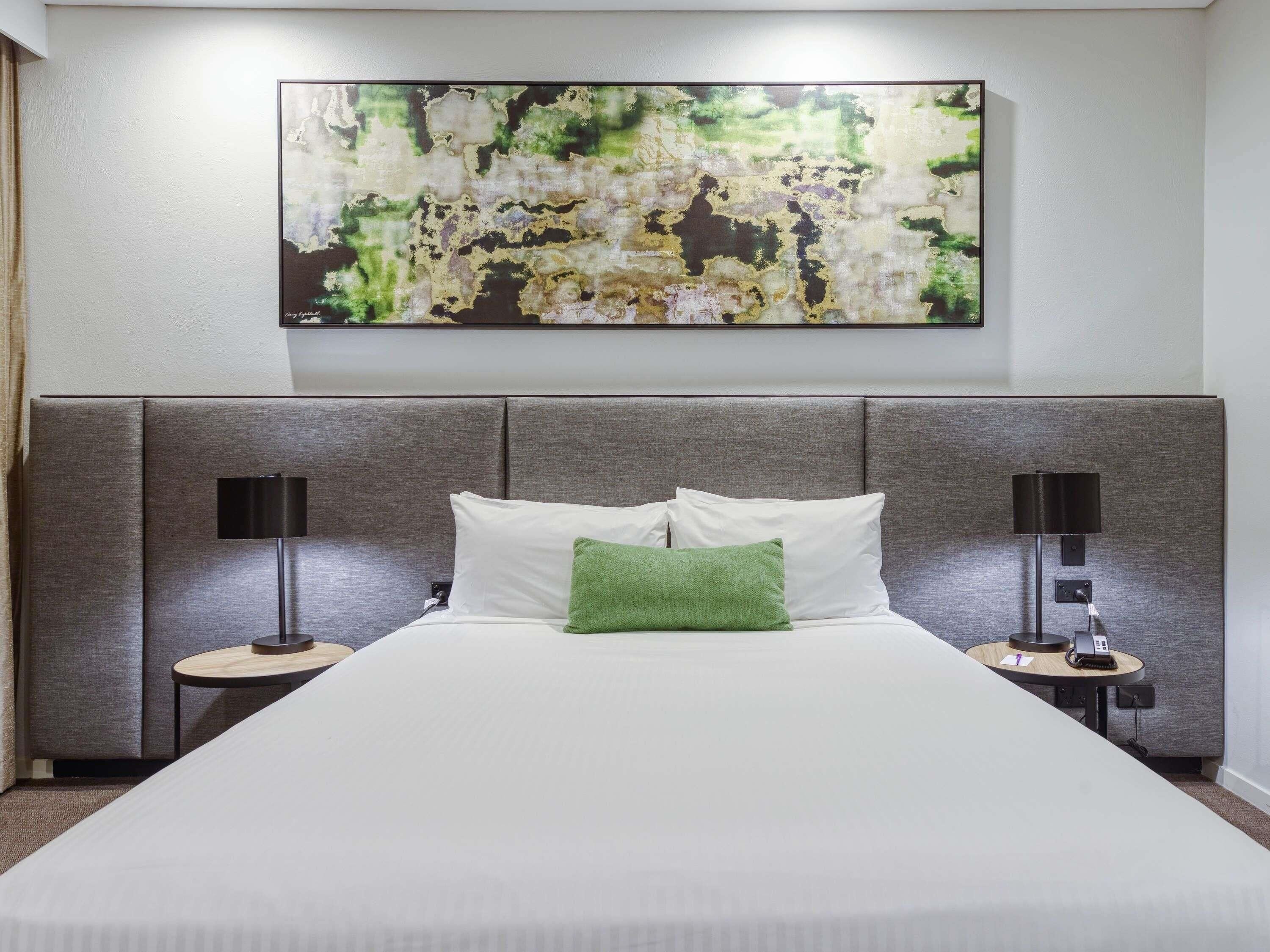 Habitación de hotel con una cama grande, dos mesitas de noche, una obra de arte en la pared y un cabecero moderno.