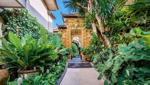 A narrow pathway leading to a traditional gate with intricate carvings, surrounded by lush greenery and potted plants. - Rama Garden Hotel (Legian)