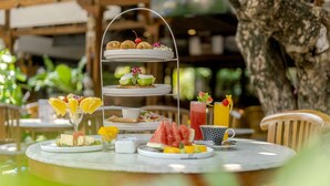 A three-tiered dessert stand with various pastries and fruits, a glass of orange juice, and a watermelon on a table with a tree in the background. - Rama Garden Hotel (Legian)