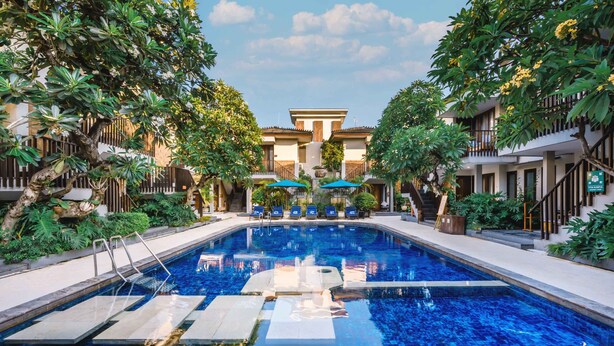 A swimming pool with clear blue water, surrounded by lush greenery and a building with a modern design. - Rama Garden Hotel (Legian)