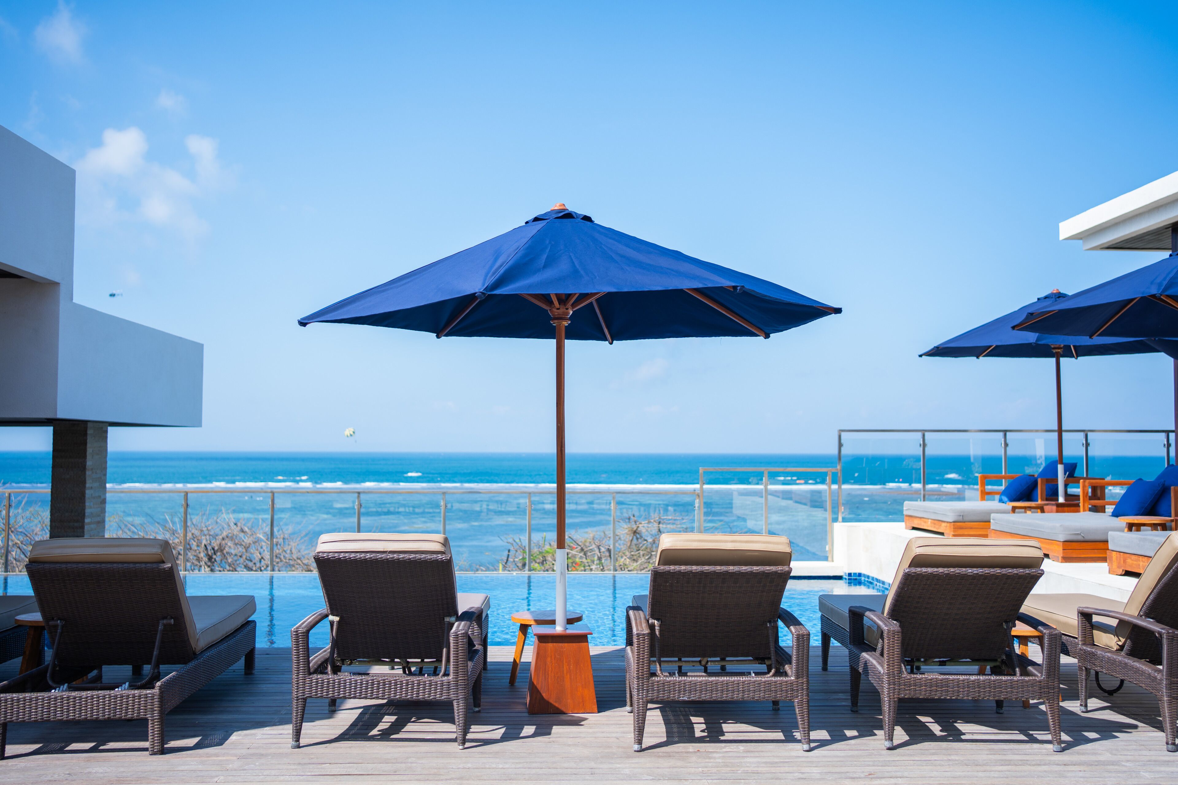Une terrasse dotée de fauteuils de jardin en osier et de parasols bleus, offrant une vue sur une piscine et l’océan.