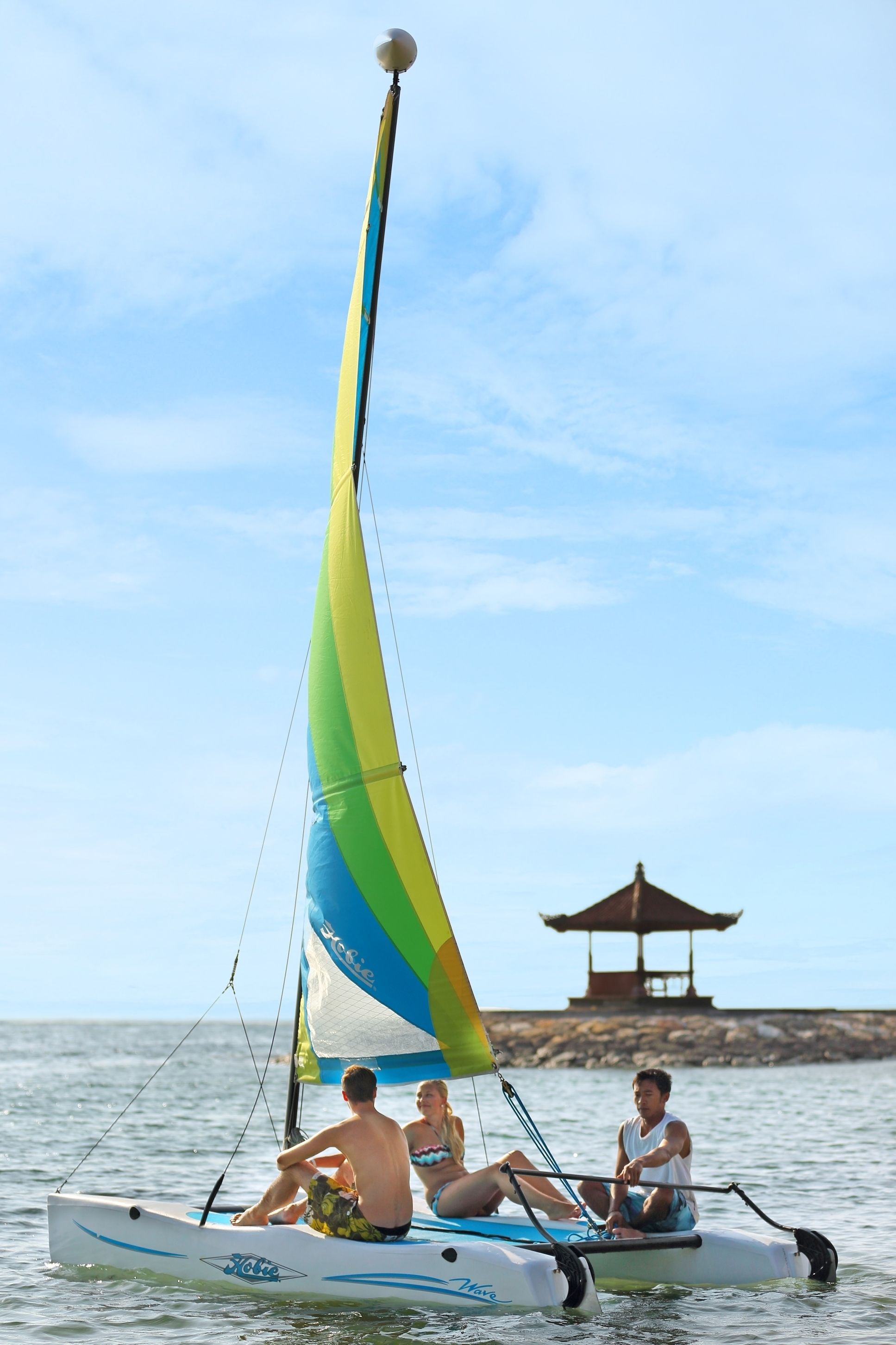 Un velero con tres personas a bordo, cielo despejado y un pabellón en el fondo.