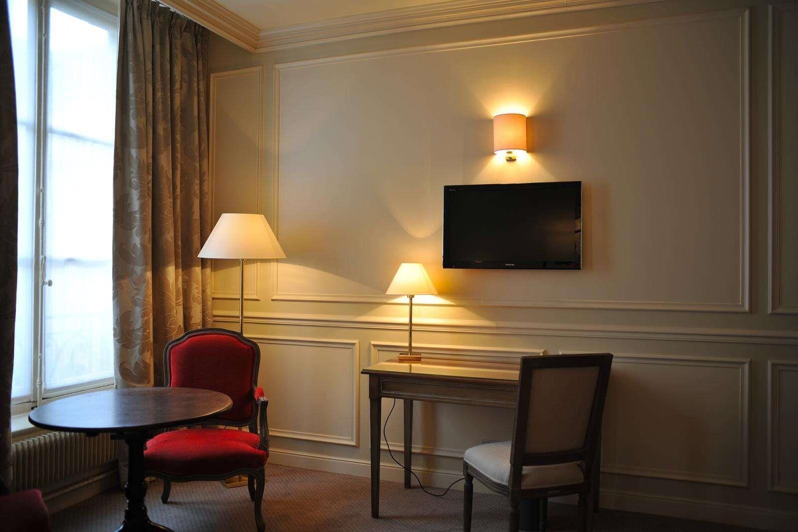 A hotel room with a red chair, a table, a TV mounted on the wall, and two floor lamps.
