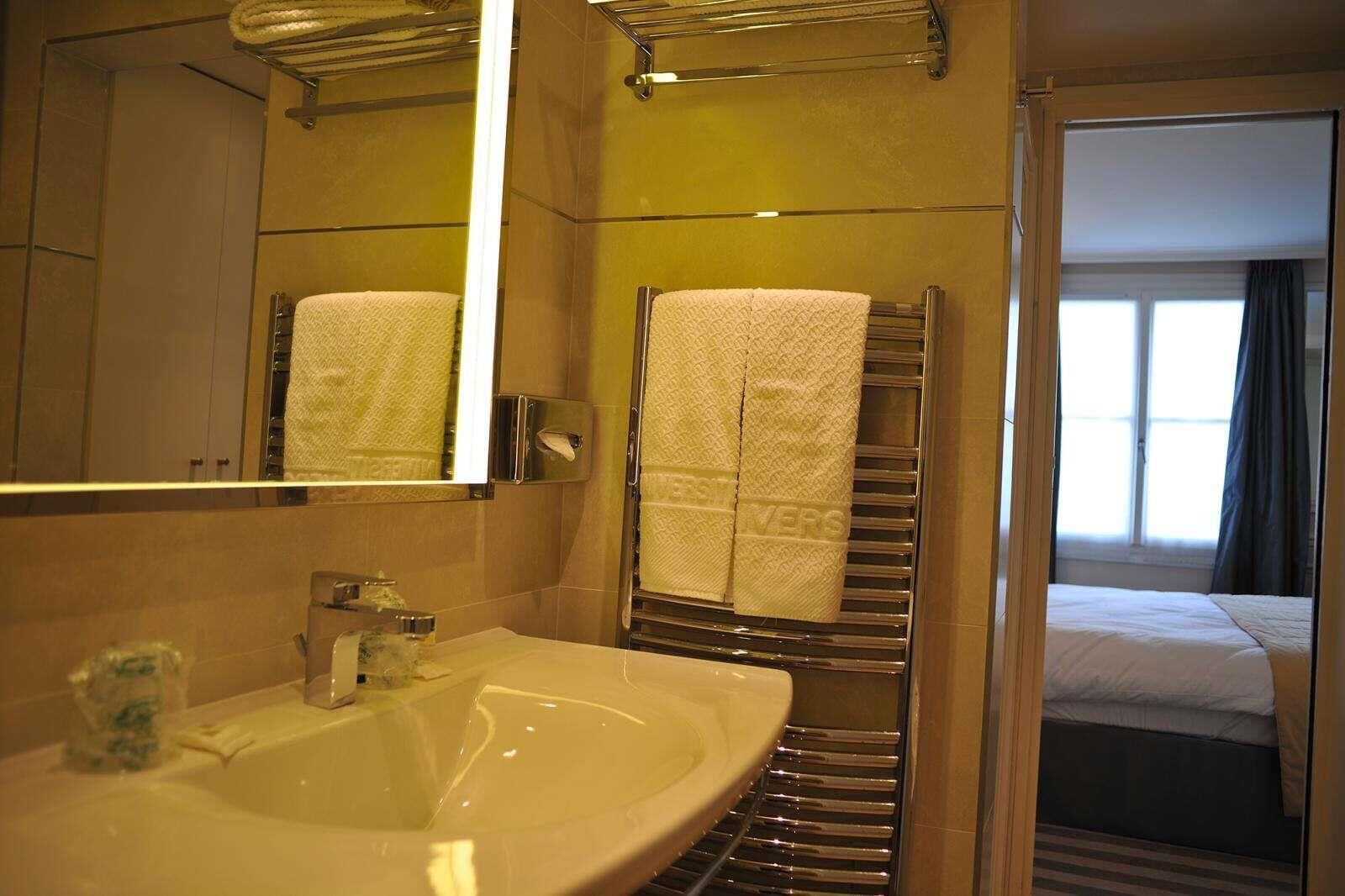 Une salle de bain d’hôtel équipée d’un lavabo, d’un miroir, d’un porte-serviettes et offrant une vue sur une chambre avec un lit et une fenêtre.