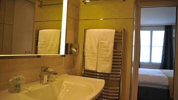 Une salle de bain d’hôtel équipée d’un lavabo, d’un miroir, d’un porte-serviettes et offrant une vue sur une chambre avec un lit et une fenêtre.