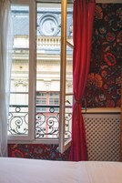 Una habitación con ventana que presenta barandales de hierro ornamentales, una cortina roja y un papel tapiz con motivos florales.