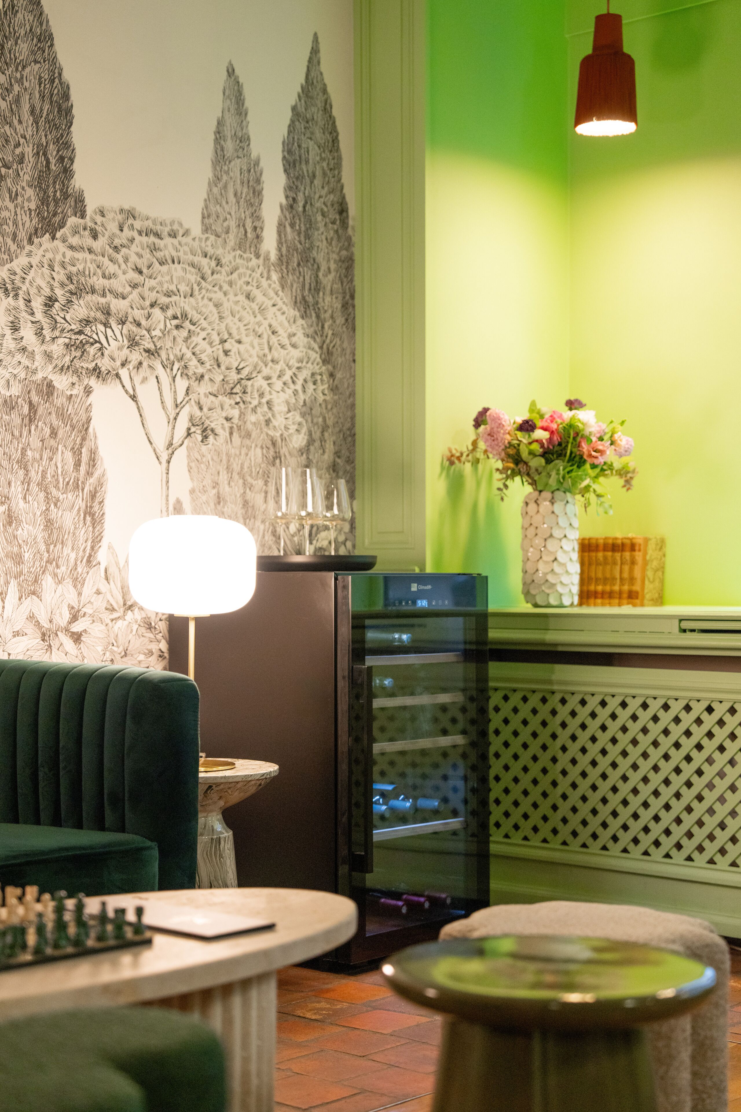 Una habitación con un sofá verde, un tablero de ajedrez, una mesa con una lámpara, un jarrón decorativo con flores y una pared con un mural de temática natural.