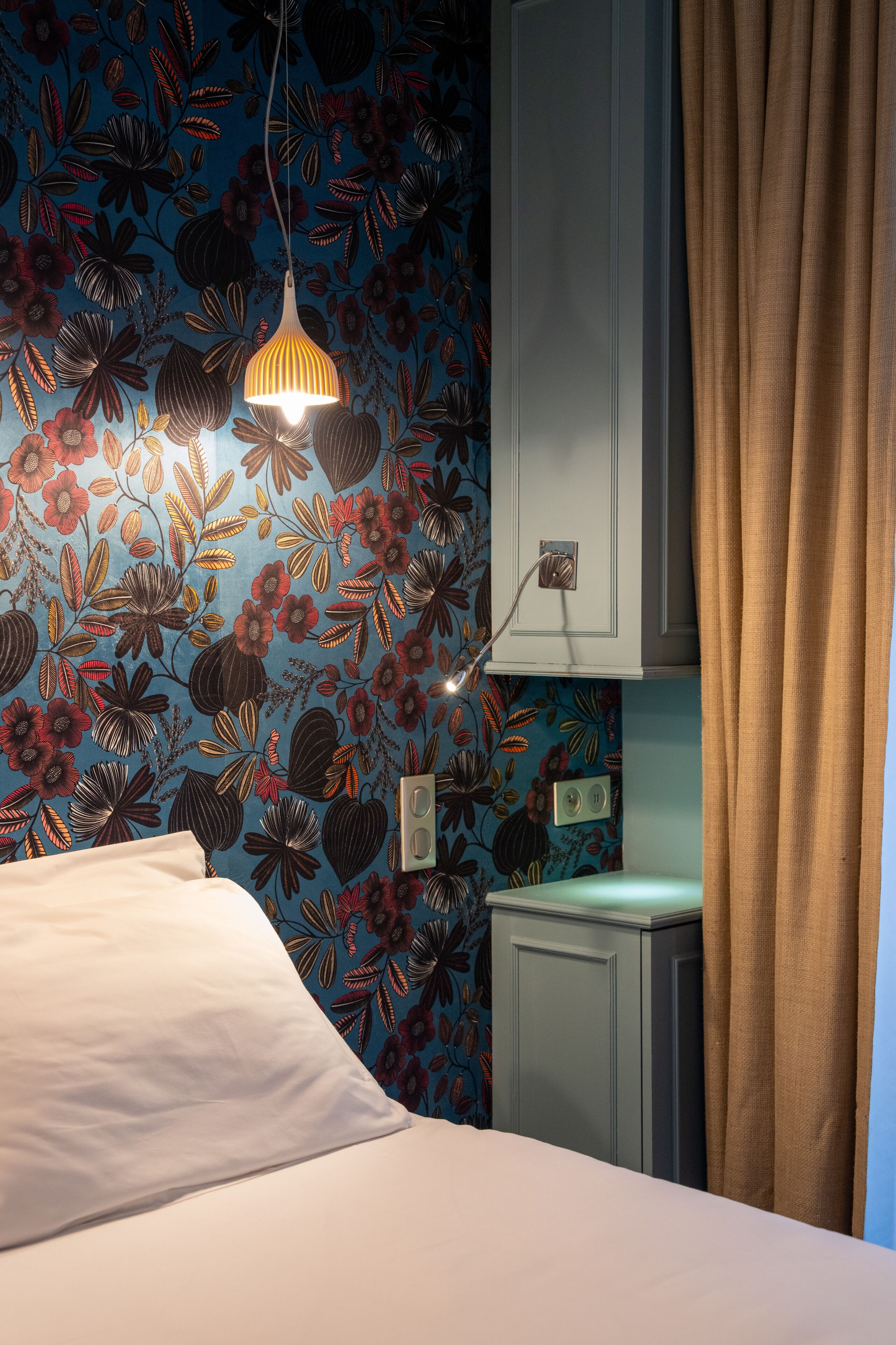 A bedroom with a floral wallpaper, a bed with white bedding, a hanging lamp, and a cabinet above the bed.