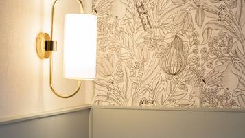 Un llit ben arropat amb roba de llit blanca, una làmpada de paret i un paper pintat amb motius florals.