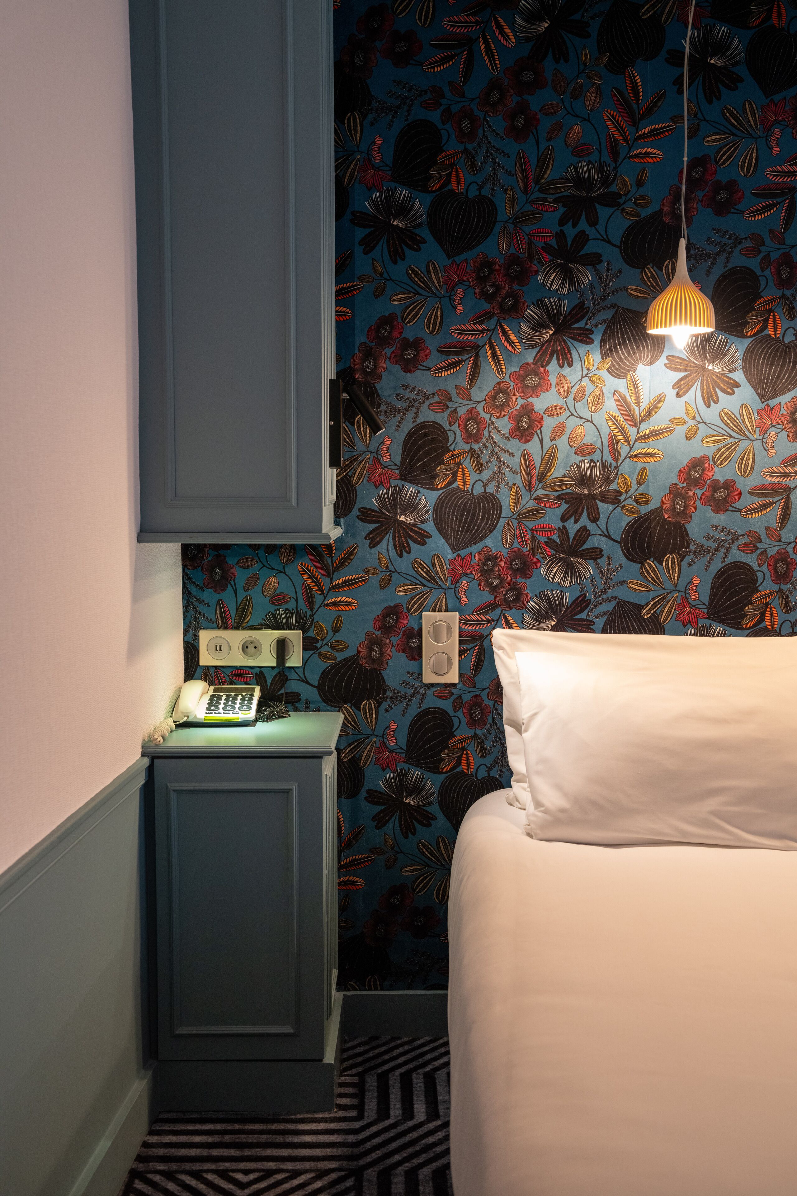 A bedroom with a floral wallpaper, a bed with white bedding, a nightstand with a telephone, and a hanging light fixture.