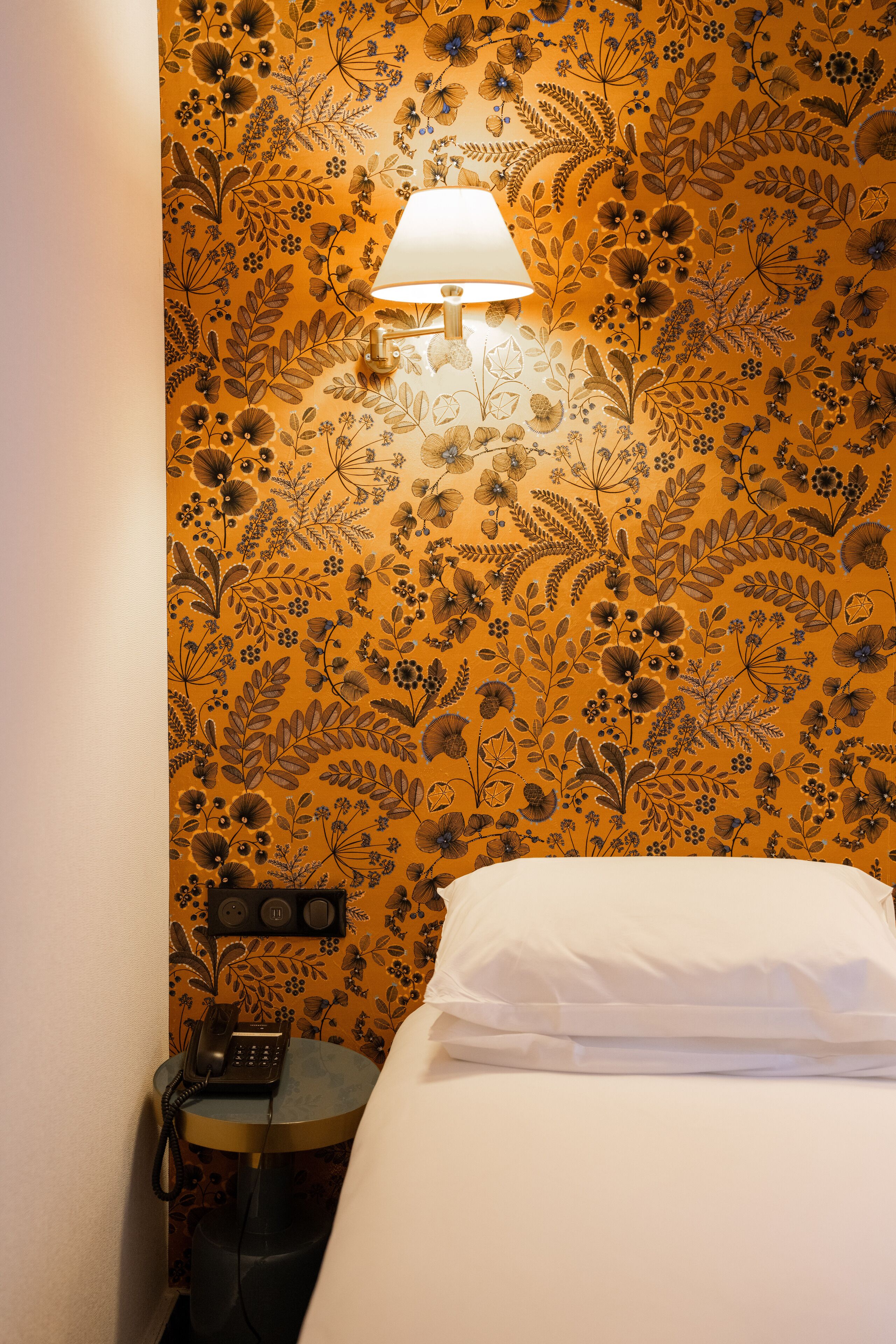 Habitación de hotel con papel tapiz floral, una lámpara de noche, un teléfono y una cama bien tendida.