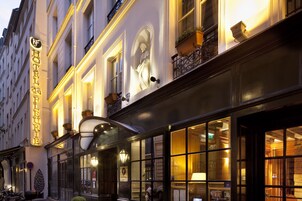 Fachada de hotel con señalización iluminada, ventanas amplias y detalles decorativos como plantas en macetas y una estatua.