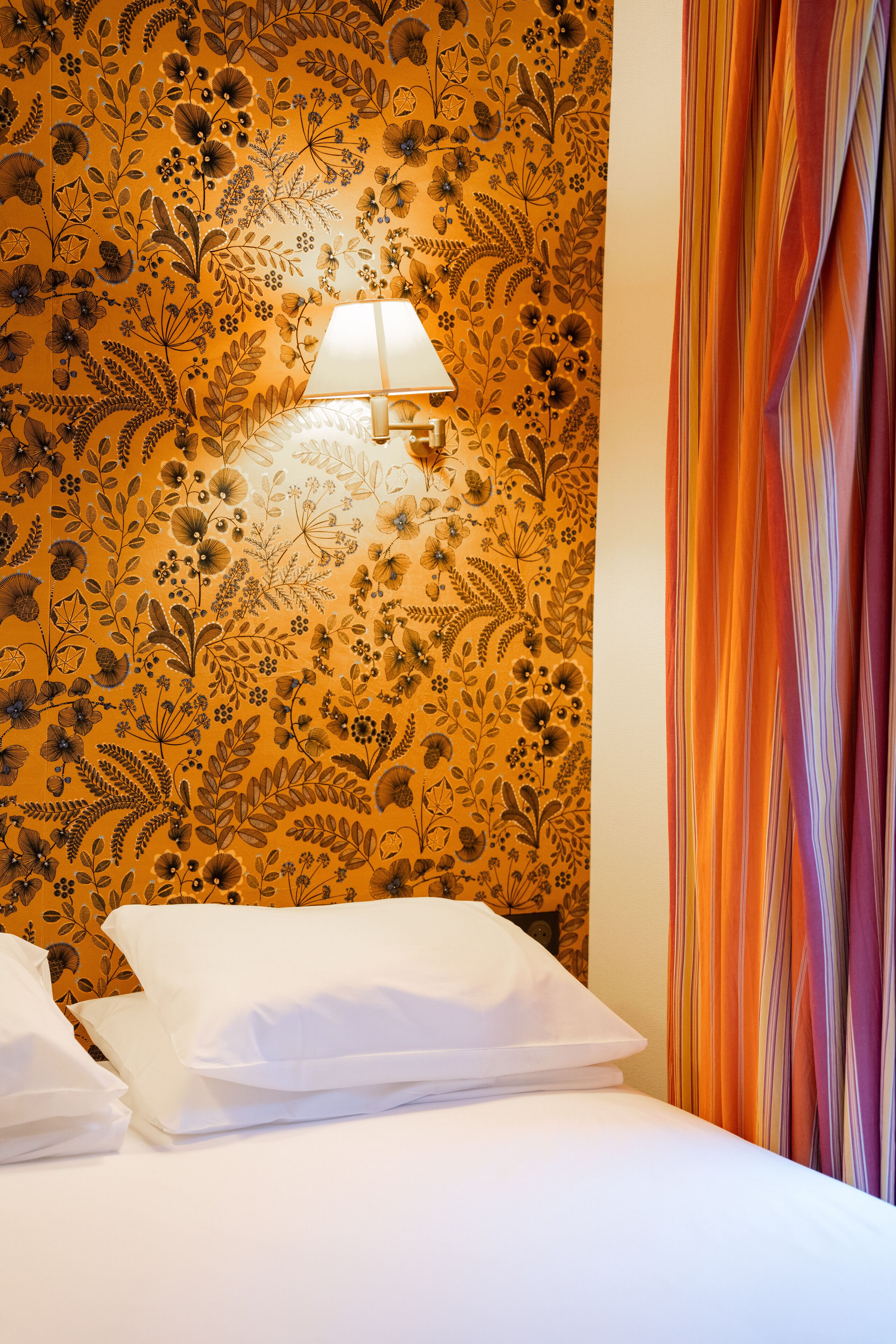 Une chambre d’hôtel avec un papier peint à motifs floraux, un lit avec du linge de maison blanc et un rideau à rayures.