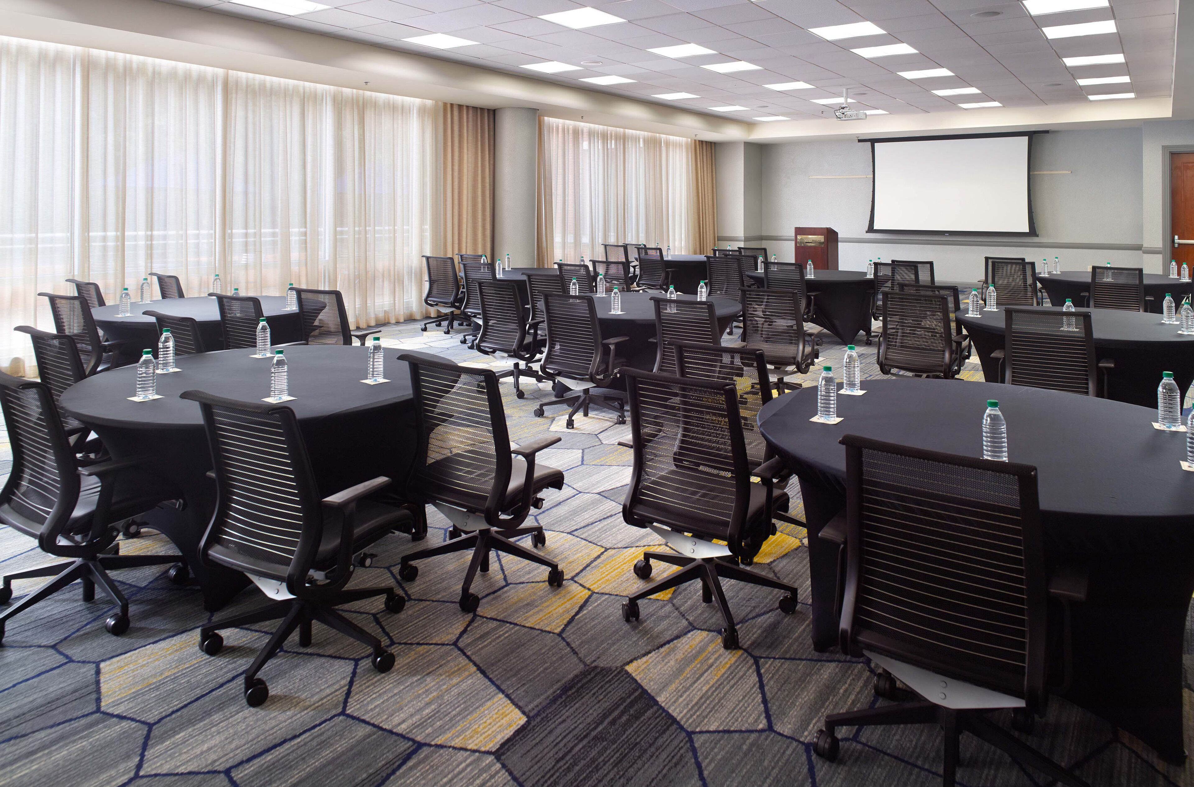 A conference room with multiple tables and chairs, equipped with water bottles, a projector screen, and a large window with curtains.
