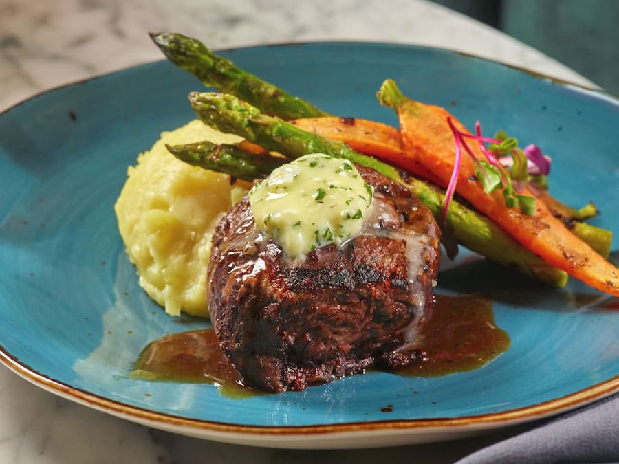 A plate with a steak topped with a creamy sauce, accompanied by mashed potatoes and roasted vegetables.