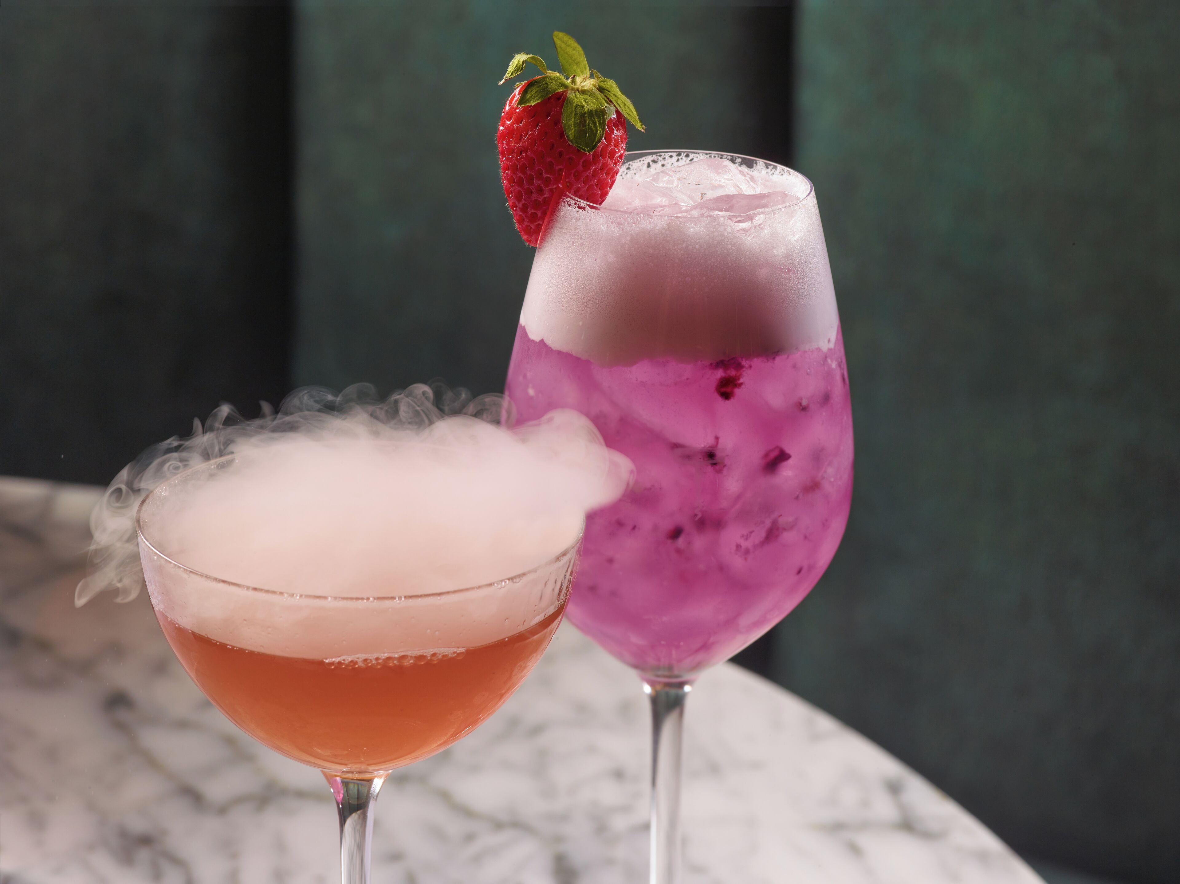 Two cocktails in glasses, one with a strawberry garnish, on a marble surface.