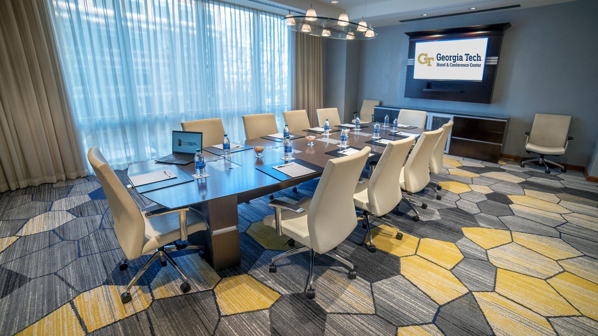 Ett konferensrum med ett stort bord, stolar, en bärbar dator, vattenflaskor och en skärm som visar texten ”Georgia Tech Hotel & Conference Center”.