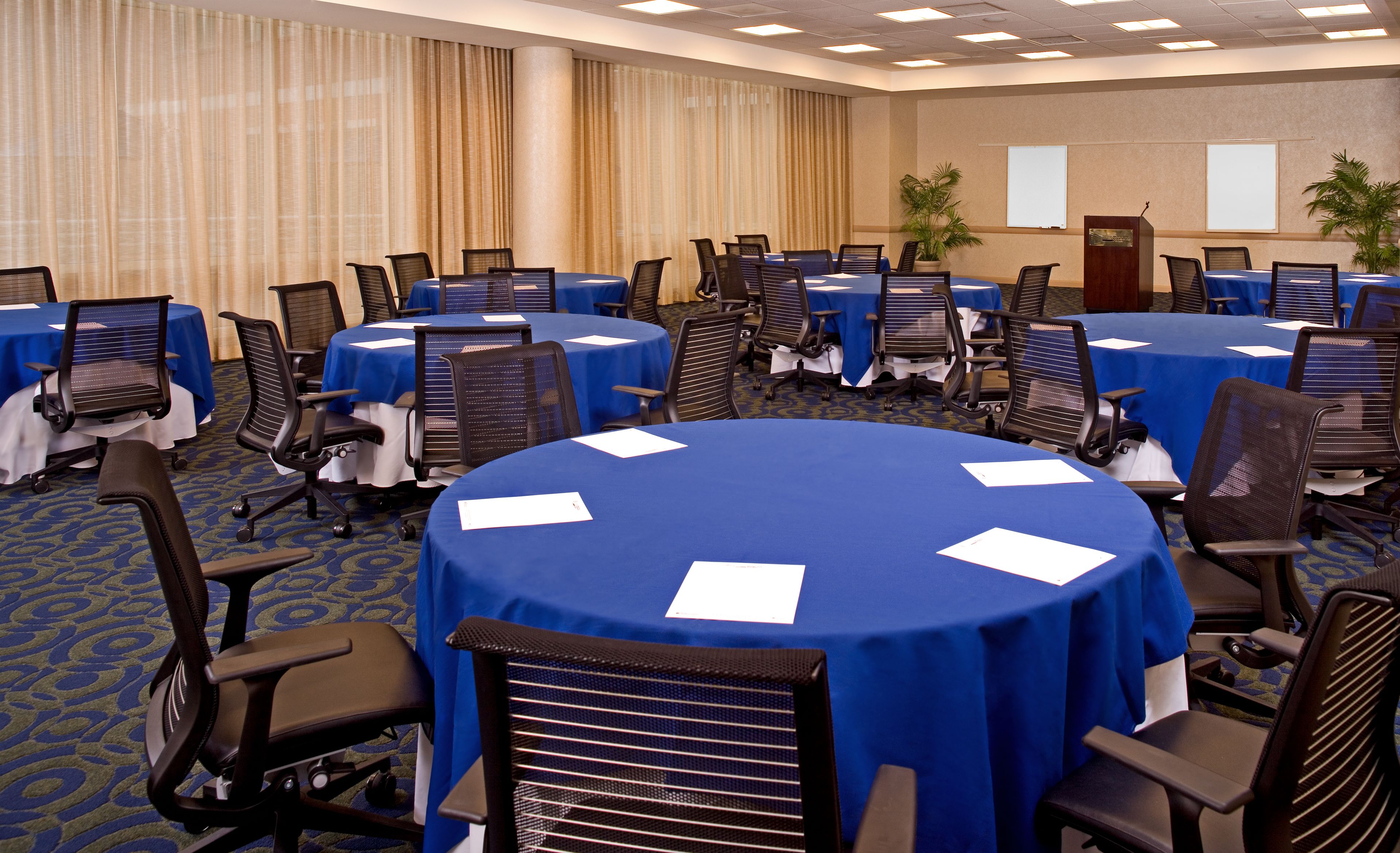 A conference room with multiple tables and chairs, arranged in a U-shape, with a projector screen and a whiteboard on the wall.