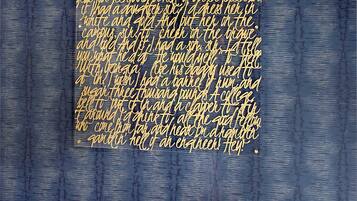 Quarto com uma grande obra de arte na parede, exibindo texto em dourado. Um banco com assento e encosto marrons está posicionado em frente à obra.