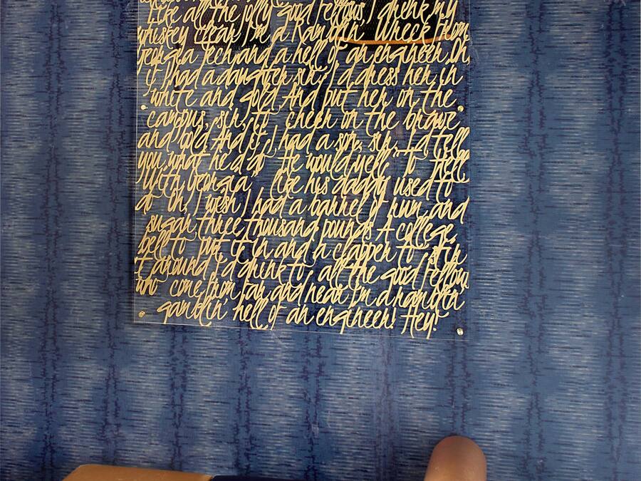 Quarto com uma grande obra de arte na parede, exibindo texto em dourado. Um banco com assento e encosto marrons está posicionado em frente à obra.