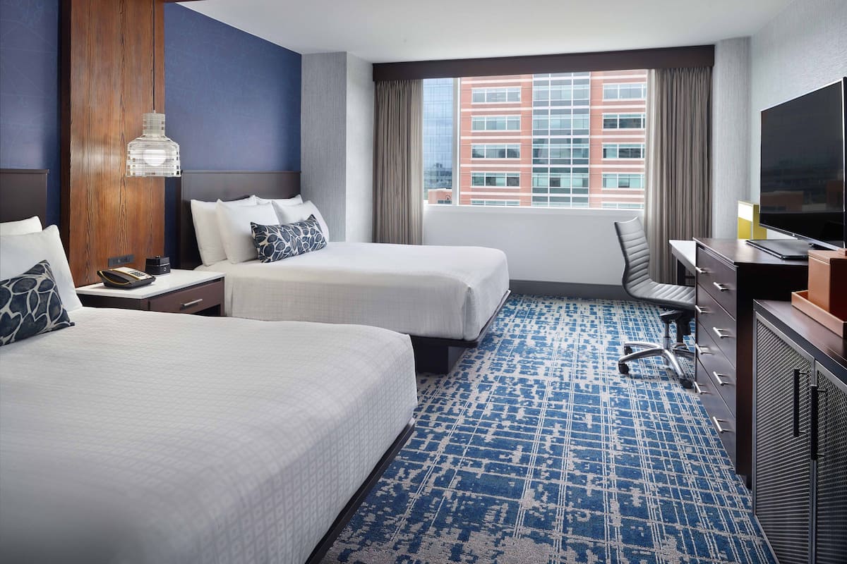 A hotel room with two beds, a desk, a chair, a TV, and a large window with a city view.