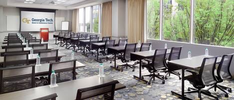 A Georgia Tech Hotel & Conference Center conference room with multiple tables and chairs, large windows with a view of trees, and a presentation screen.