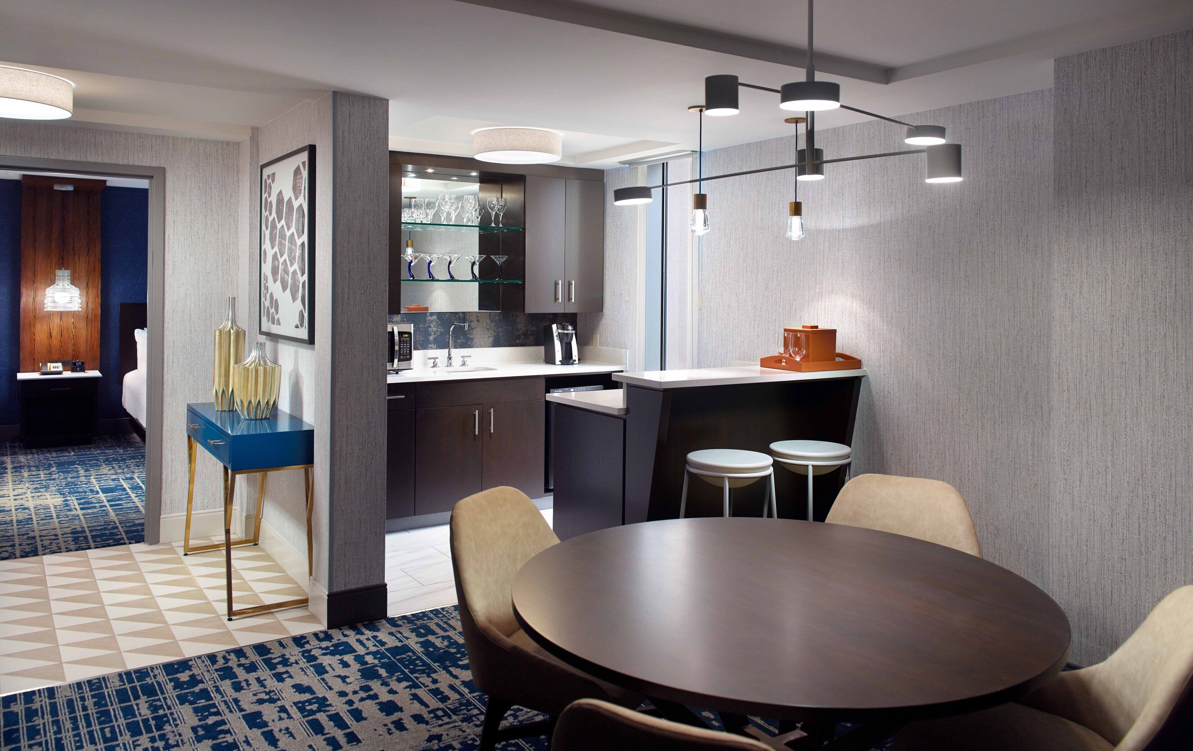 A modern hotel room with a dining area, a kitchen, and a bathroom.