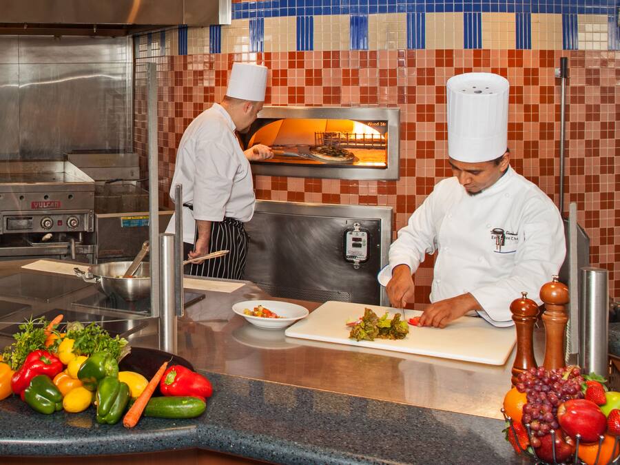Une cuisine professionnelle où des chefs préparent des plats, avec un four à bois et une variété de légumes frais disposés sur le plan de travail.